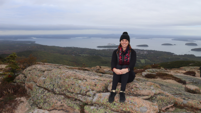 Phoenix College alumna Chelsie Parish sits on top of a mountain with a wide open landscape of greenery and waterway behind her.    