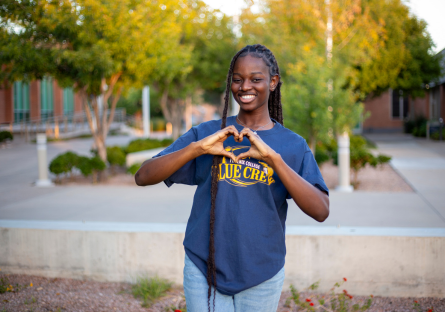 Fall 2024 graduate Mimi Owolabi wears a Phoenix College t-shirt and holds her hands in a heart shape while standing on campus