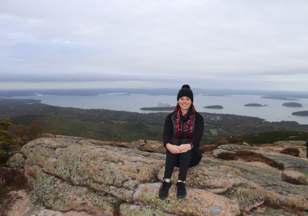 Phoenix College alumna Chelsie Parish sits on top of a mountain with a wide open landscape of greenery and waterway behind her.    