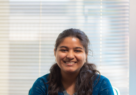 PC student Keerthie Veluri sits in front of white window blinds smiling; she is prepared to graduate with a bachelor's in IT by the time she is 18 years old. 