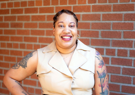 Pamela Smith stands smiling in front of a red brick wall wearing a beige sleeveless coat dress with her tattooed arms visible and one hand on her hip