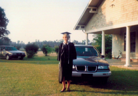 Kimberly Britt standing outside in a cap and gown next to a car at her foster home