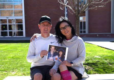 Phoenix College Alumni Jae Staats and Nancy Chong sit on a bench outside Bullpitt Auditorium, with the Phoenix College library in the background, holding a picture of themselves at their 1993 PC graduation