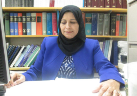 PC engineering professor Dr. Amal Saeed Yagub sits at her desk looking at papers on her desk with a bookcase of books behind her