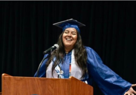 Phoenix College graduate Ivonne Dominguez in her cap and gown at a podium speaking with her arms outstretched