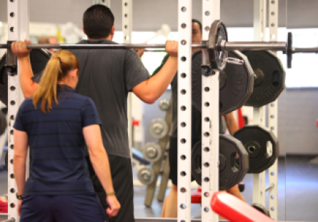 A man performs weighted squats on a Smith machine supervised by a female fitness coach in Phoenix College's Fitness Center 