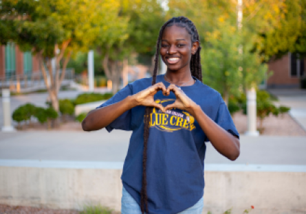 Fall 2024 graduate Mimi Owolabi wears a Phoenix College t-shirt and holds her hands in a heart shape while standing on campus