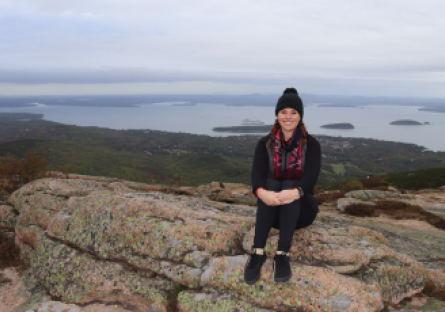 Phoenix College alumna Chelsie Parish sits on top of a mountain with a wide open landscape of greenery and waterway behind her.    