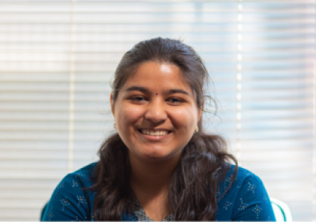 PC student Keerthie Veluri sits in front of white window blinds smiling; she is prepared to graduate with a bachelor's in IT by the time she is 18 years old. 