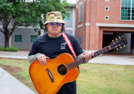 Musician and PC songwriting student Troy Lowney stands on campus outside the recording studio wearing a black t-shirt, sunglasses and a camouflage hat with a guitar strapped over his shou